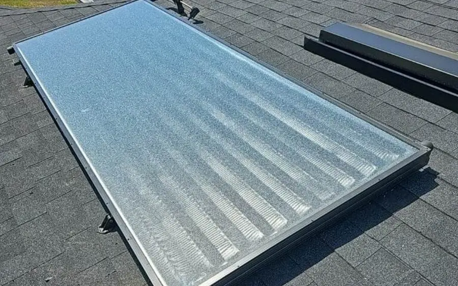 A glazed solar water heating collector mounted flat on a shingle roof, with its supply and return plumbing visible at the lower corner.